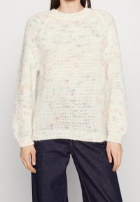 Personne portant un pull en maille lâche crème avec des taches multicolores, associé à un jean bleu foncé à jambes larges, visible du cou aux hanches.