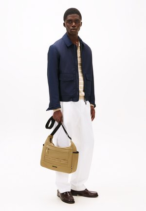 Giacca blu navy, camicia a righe beige e bianca, pantaloni bianchi, scarpe marroni, con una borsa a tracolla beige con cerniere e tasche.