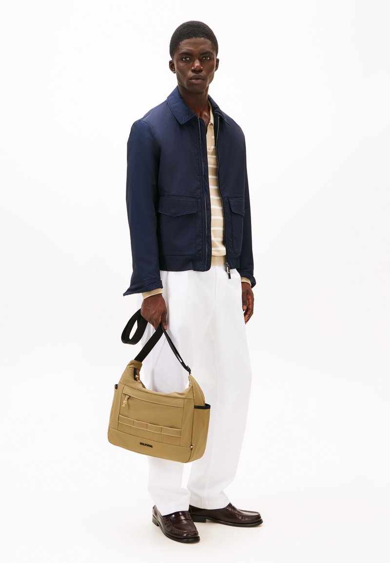 Navy jacket, striped beige and white shirt, white trousers, brown shoes, holding a beige shoulder bag with zippers and pockets.