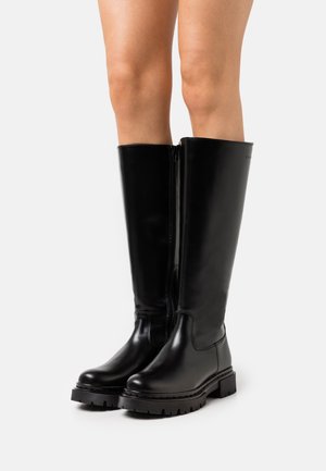 Jambes portant des bottes en cuir noires montant jusqu'aux genoux avec des semelles épaisses à crampons et des fermetures éclair sur le côté intérieur, sur fond blanc.