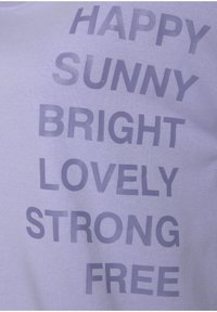 Camiseta lila con gran texto en tamaños variados: "HAPPY," "SUNNY," "BRIGHT," "LOVELY," "STRONG," y "FREE," en letras impresas de color púrpura oscuro.