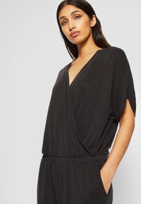 Svart, omslags jumpsuit med korte ermer, V-hals og elastisk midje. Mykt, lett stoff med en jevn tekstur.