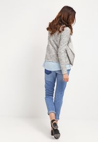 Grå texturerad jacka med draperad design, blå skjorta under, i kombination med ljusblå jeans och svarta klackskor.