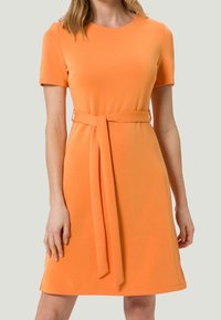 Pomarańczowa sukienka krótko rękawowa z okrągłym dekoltem i wiązaniem w pasie. Materiał wydaje się gładki, a krój jest dopasowany w górnej części, rozszerzający się przy dole.