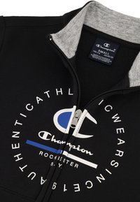 Chaqueta negra con cremallera y cuello gris, que presenta un gran logo circular blanco y azul junto con texto. El material parece suave con un interior blando.