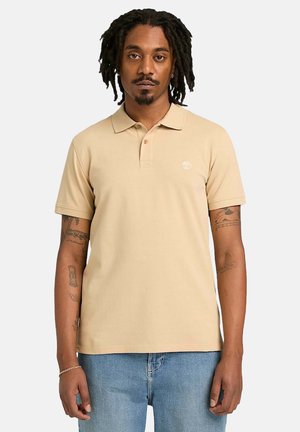 Uomo con dreadlocks che indossa una polo beige a maniche corte e jeans azzurri, in piedi davanti a uno sfondo bianco uniforme.