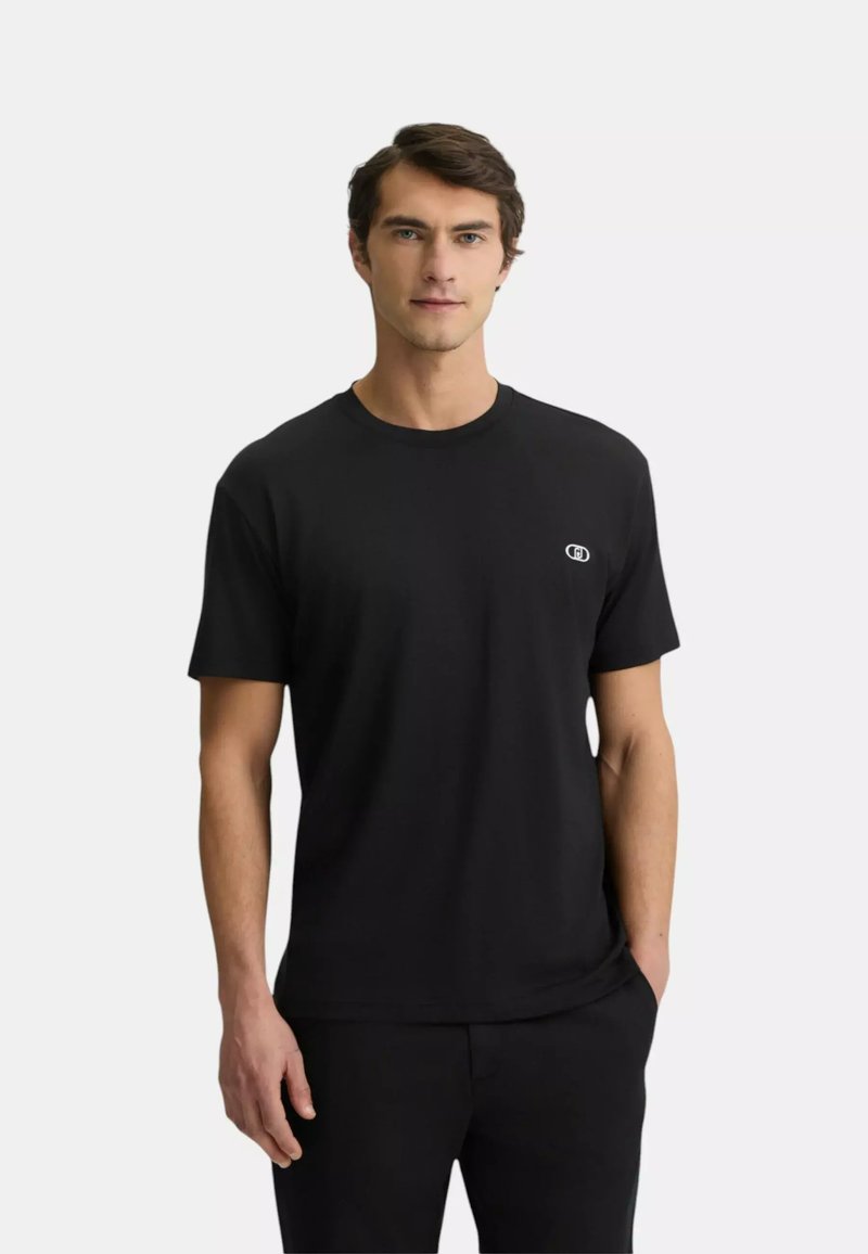Homme portant un t-shirt noir uni à manches courtes avec un petit logo blanc sur le côté gauche de la poitrine, debout devant un fond clair.