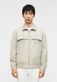 Ανοιχτό μπεζ μπουφάν με κλείσιμο με φερμουάρ, ελαστικά μανίκια και ελαφρώς φαρδιά εφαρμογή. Διαθέτει οριζόντιο μπροστινό πάνελ και πλαϊνές τσέπες.