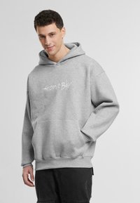 Siv Hoodies s sprednjo žepom, ki vsebuje bel napis "Home Boy". Oblika vključuje prostoren kapuco in rebraste manšete.