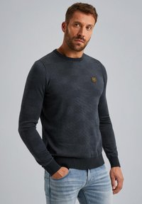 Tmavošedý sveter s textúrovaným pleteným vzorom, okrúhlym výstrihom a rebrovanými manžetami. Obsahuje malú zlatú nášivku s logom na hrudi.