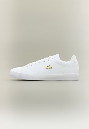 Baskets basses blanches avec des lacets blancs, une semelle en caoutchouc plate et un logo crocodile doré sur le côté, sur un fond neutre.