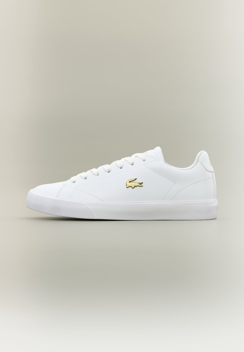 Baskets basses blanches avec des lacets blancs, une semelle en caoutchouc plate et un logo crocodile doré sur le côté, sur un fond neutre.