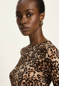 Frau mit kurzen geflochtenen Haaren, die goldene Ohringe in Form von Ringen trägt und ein langärmeliges Oberteil mit Leopardenmuster anhat, schaut leicht zur Seite.