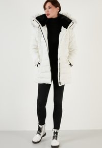 Manteau doudoune blanc avec une capuche en fausse fourrure noire, fermetures éclair et pressions, et poches latérales, associé à un legging noir et des bottes blanches à lacets.