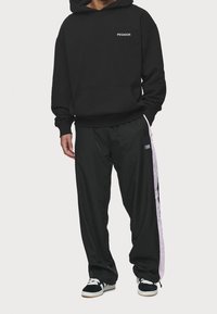 Felpa nera con cappuccio decorata con il logo "PEGADOR", abbinata a pantaloni neri larghi con una striscia rosa laterale e sneakers nere.