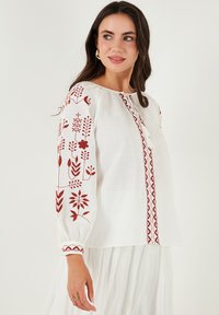 Blouse en lin blanc ornée de broderies florales rouges sur les manches et le devant, avec des poignets froncés et un détail de col à boutons.