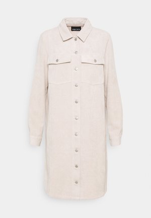 Beige corduroy overhemdjurk met kraag, lange mouwen, twee borstzakken en zilverkleurige knoopsluitingen aan de voorkant. Verticale geribbelde textuur.