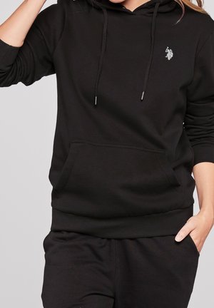 La sudadera negra cuenta con un bolsillo frontal, capucha con cordones y un logo bordado en el pecho. Está hecha de un suave tejido texturizado con puños acanalados.