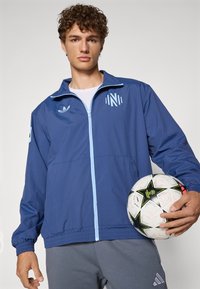 Blå lettvektsjakke med hel glidelås, med lyseblå detaljer og logoer. Modellen holder en svart-hvitt fotball.