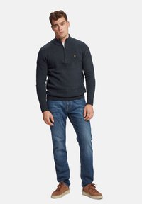 Temno siva zip-up jopica z rebrastim ovratnikom in robom, oblečena čez modre kavbojke in rjave superge, z majhnim logotipom na prsih.