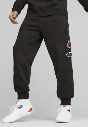 Person wearing black track pants with colorful patches and white sneakers with red and blue accents, standing against a gray background.