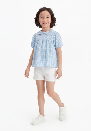 Νεαρή κοπέλα που χαμογελά, περπατώντας, φορώντας ανοιχτό μπλε καρό μπλούζα με γιακά Πίτερ Παν, λευκά σορτς και λευκά αθλητικά παπούτσια σε λευκό φόντο.