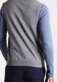 Pull sans manches gris avec un col rond, porté sur une chemise bleue à manches longues ; texture lisse et détail de bord-côte à l'ourlet.