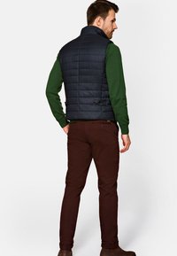 Granatowa kamizelka puchowa z wysokim kołnierzem, pikowana, z pięcioma guzikami z tyłu, zestawiona z ciemnozielonym swetrem i brązowymi spodniami.