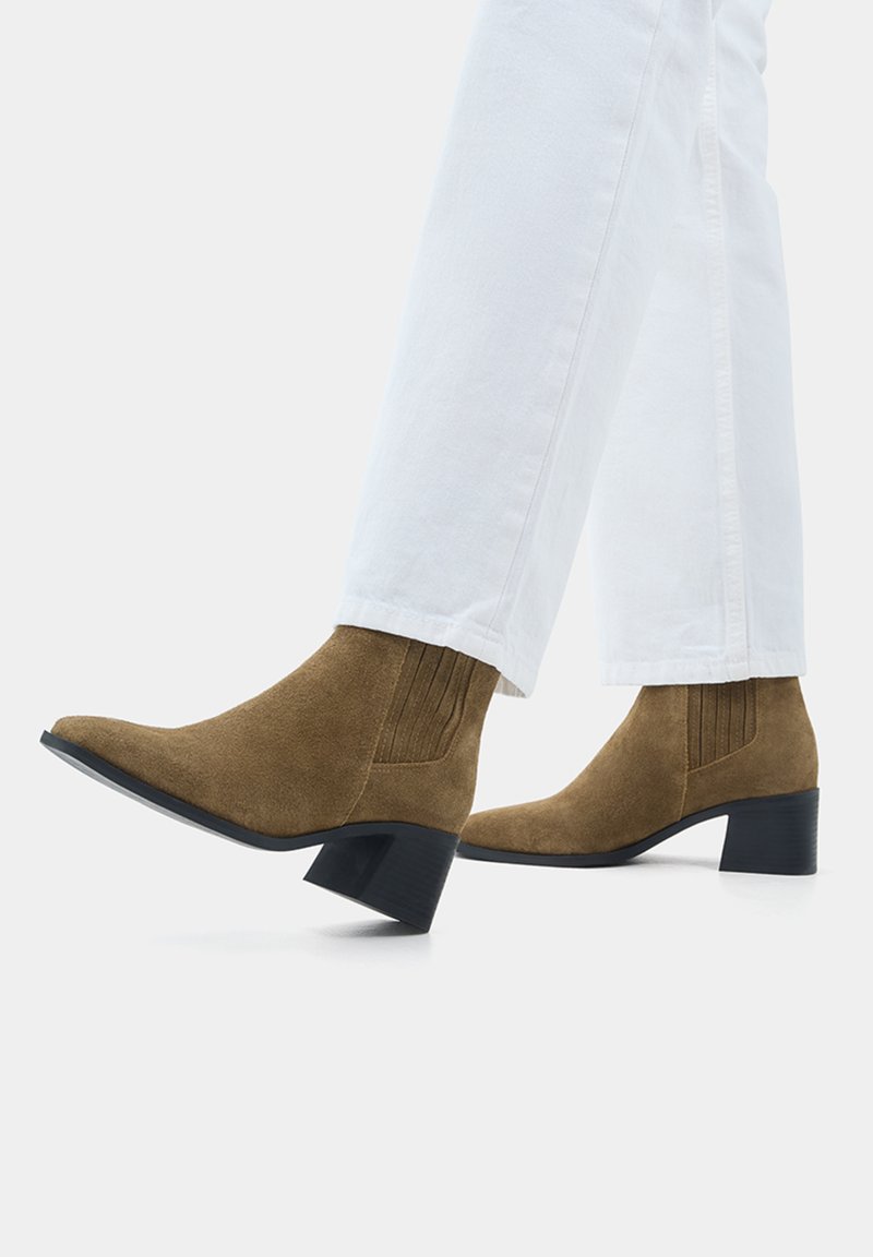 Bottes chevilles en suede marron avec un bout pointu et un talon bloc de 5 cm. Comprend des panneaux élastiques latéraux pour un enfilage plus facile et une semelle lisse noire.