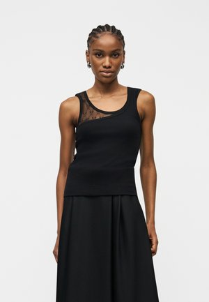 Fonott hajú nő, aki ujjatlan fekete felsőt visel, áttetsző virágos betéttel és fekete szoknyával, fehér háttér előtt áll.