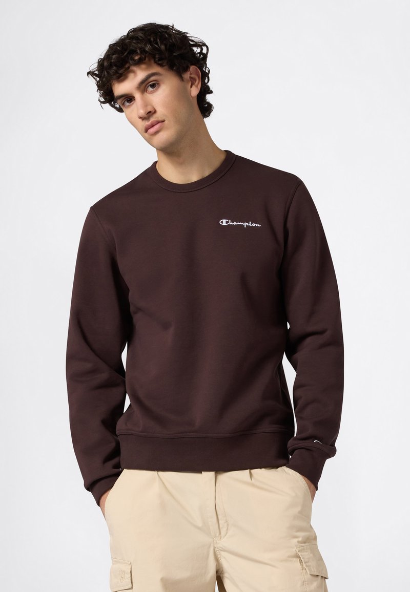 Giovane con capelli ricci che indossa una felpa Champion marrone scuro e pantaloni cargo beige, in piedi con le mani in tasca su uno sfondo bianco.