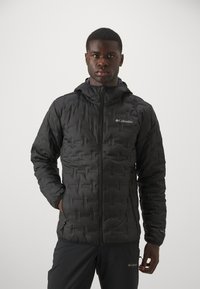 Veste matelassée noire avec une fermeture éclair à l'avant et une capuche. Elle présente un motif texturé, avec le logo Columbia sur la poitrine et des poches zippées.