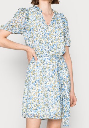 Vrouw die een korte, ceintuurloze bloemenjurk met geplooide mouwen draagt in blauw-, groen- en beige tinten tegen een effen achtergrond.