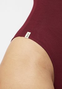 Bodysuit bordeaux avec une texture lisse, doté d'une petite étiquette en crème à la ceinture. Design simple et ajusté.