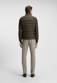 Braune Steppweste mit hohem Kragen und gestepptem Muster, getragen über einem Langarmhemd und hellgrauen Hosen. Elegantes Design, glatte Textur.