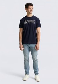 Laivastonsininen puuvillainen t-paita, jossa painettu logo ja teksti rinnan kohdalla, yhdistetty vaaleansinisiin farkkuihin ja valkoisiin lenkkareihin.