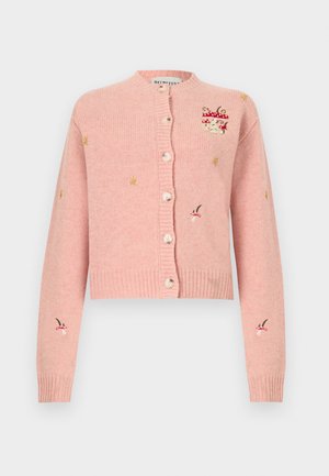 Femeie purtând un cardigan roz brodat, blugi largi albaștri și mocasini negri, stând în picioare pe un fundal simplu și deschis la culoare.