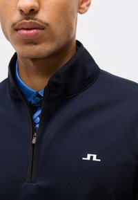 Κοντινό πλάνο ενός ατόμου που φοράει ένα μπλε μπουφάν με φερμουάρ πάνω από ένα μπλε πουκάμισο με γιακά, με ένα μικρό λευκό λογότυπο στο στήθος του μπουφάν.