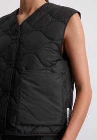 Zwarte gewatteerde gilet met een V-hals, mouwloos ontwerp, voorzak met klep en logo-label aan de zijkant. Glad, lichtgewicht stof.