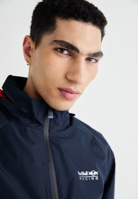 Mornarski windbreaker s visokom kragnom, s patentnim zatvaračem i logotipom Red Bull Racing. Glatka tekstura s podesivim detaljima.