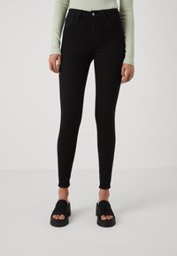Woman wearing black skinny jeans, a light green ribbed long-sleeve top, and black open-toe platform sandals on a neutral background.