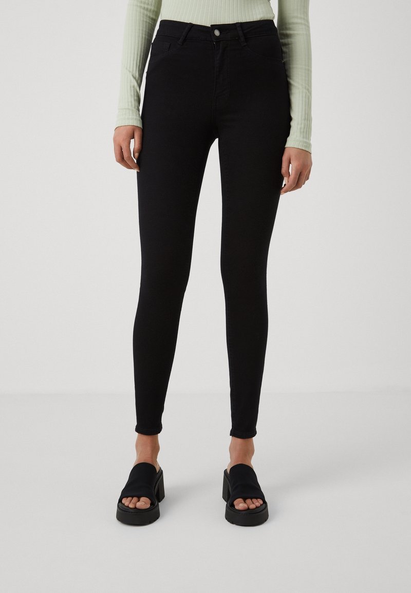 Woman wearing black skinny jeans, a light green ribbed long-sleeve top, and black open-toe platform sandals on a neutral background.