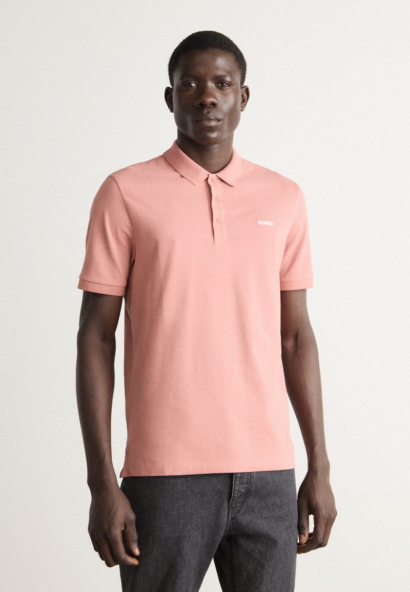 Polo rose clair en coton avec un col, trois boutons et un petit logo blanc « HUGO » sur la poitrine gauche.