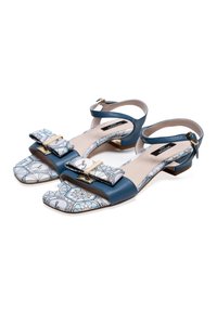 Sandales en cuir bleu avec une lanière ornée de motifs, dotées d'un nœud décoratif et de quincaillerie dorée. Talon bas et bride de cheville réglable.