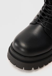 Juodos odinės kulkšnės botas su apvaliu pirštu, storu guminio sulankstyto krašto padu ir tekstūruota paviršiumi. Šniūrelės tęsiasi iki vidurinės kojinės.