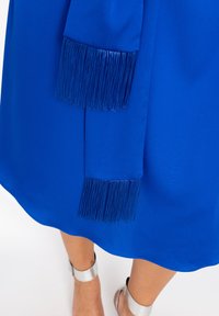 Blauwe jurk met een gladde stof en een golvende zoom. De mouwen hebben een horizontale franjesdetail en de draagster heeft zilveren enkelbandjes.