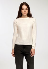 Kremowy sweter z dzianiny z długimi rękawami, okrągłym dekoltem i ściągaczami. Noszony z ciemnymi, teksturowanymi spodniami, podkreślającymi dopasowaną sylwetkę.