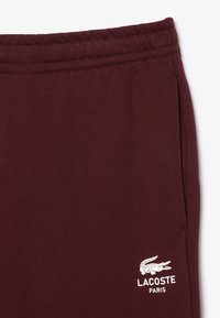 Vinröda mjukisbyxor i mjukt tyg, med elastisk midja, sidofickor och en vit Lacoste-logotyp med en krokodil-design.