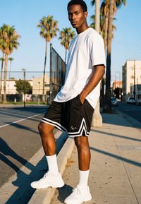 Noor mees valges T-särgis, mustades spordipükstes ja valgetes tossudes, seismas päikeselisel kõnniteel aiaga piiratud korvpalliväljaku lähedal, palmiokstega.