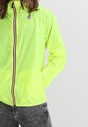 Osoba ubrana w jasną żółtą kurtkę K-Way z wielokolorowym paskowym zamkiem błyskawicznym oraz szare jeansy, stojąca z jedną ręką w kieszeni.